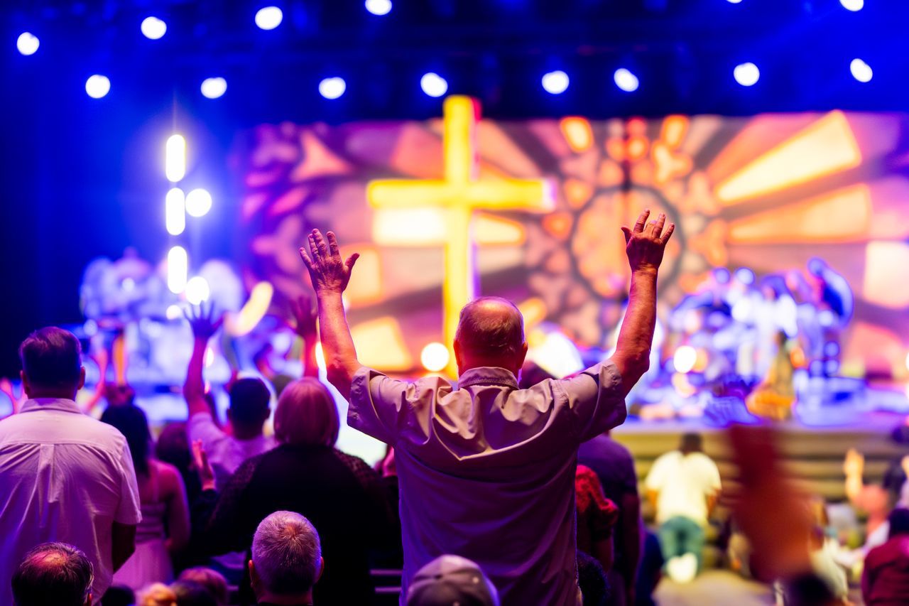 A lively worship service with people praising, colorful lights, and a cross stage backdrop.
